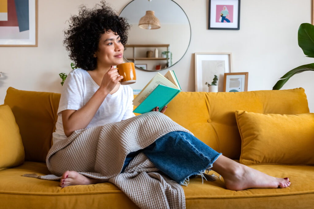 A person on a couch in a blanket, with a mug in one hand and a book in the other.