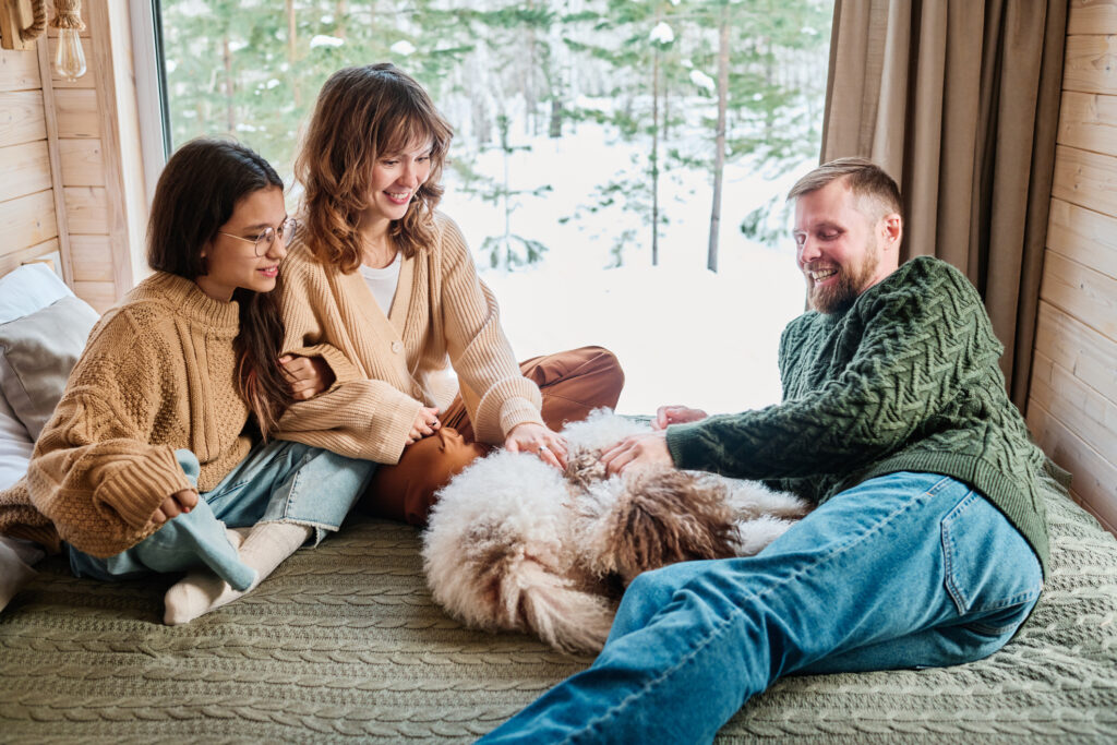 A group of friends relaxing on a bed with a dog, with a snowy scene out the window in the background.